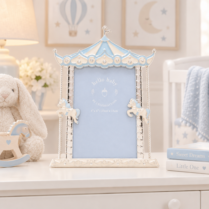 A decorative baby photo frame styled like a carousel, featuring a light blue central panel and pale blue trim with cream-colored carousel details. The frame is displayed on a white dresser alongside a plush bunny toy, books, and a soft crib blanket in a nursery setting.