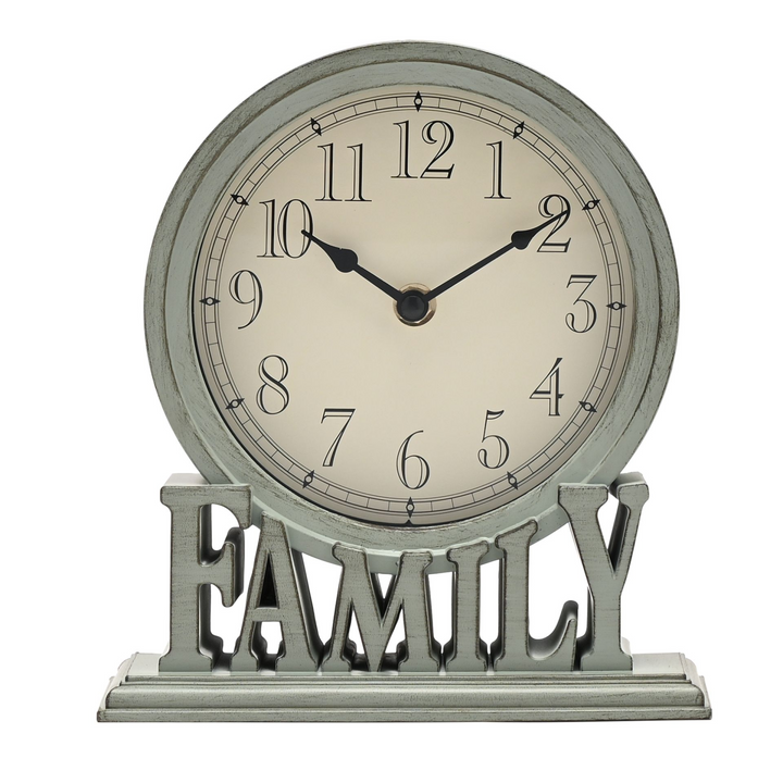 A round wall clock with a distressed sage green frame and base, featuring a cream-colored face and black numerals. The clock is mounted on a matching distressed metal stand with the word &