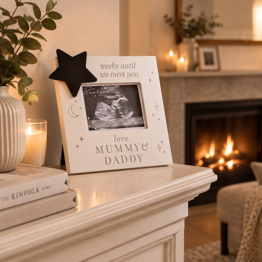 A white rectangular photo frame with black text reading 'weeks until we meet you' and 'love MUMMY & DADDY,' plus small moon and star illustrations, displayed on a fireplace mantel. The frame holds a black-and-white ultrasound photo and is styled with a black star topper, candles, books, and a cozy living-room background.
